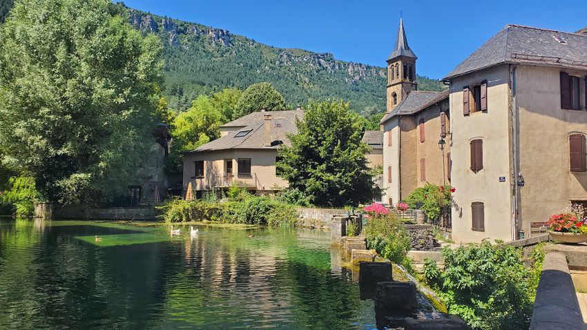 des maisons et un clocher au bord d'une retenue d'eau, un front montagneux couvert d'arbres à l'horizon