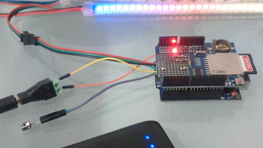 an sd card is plugged into the arduino shield, which is in turn connected by jump wires to the rainbow-lit led strip, the infrared receiver, and the power bank